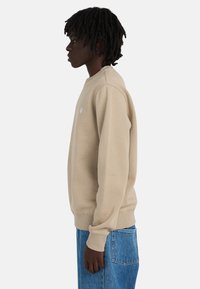 Side profile of a person with short dreadlocks wearing a beige sweatshirt and blue denim pants against a plain background.