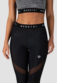 Zwarte actieve leggings met doorzichtige meshpanelen, hoge tailleband en een aansluitend ontwerp. Voorzien van een wit logo aan de voorkant. Zacht, rekbaar materiaal.