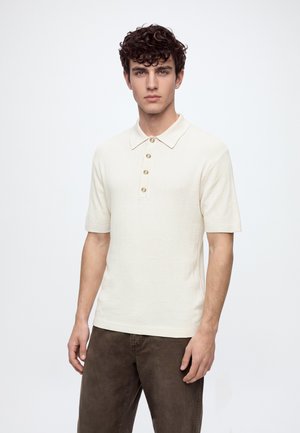 Jeune homme aux cheveux bouclés foncés, portant un polo crème à manches courtes et un pantalon marron, debout devant un fond blanc uni.