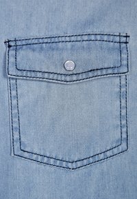 Jasnoniebieski denim z prostokątną kieszenią na piersi, wyposażoną w srebrny zatrzask i kontrastujące ciemne przeszycia wzdłuż krawędzi.