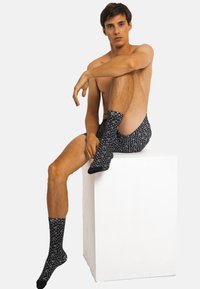 Chaussettes noires à motifs avec des designs blancs, portées avec un short noir à motifs, sur un modèle assis contre un fond blanc uni.