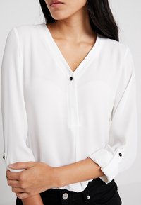 Bílá blůza s výstřihem do V a dlouhými rukávy, s černým knoflíkovým zapínáním a ohrnutými manžetami, vyrobená z hladké, lehké látky.