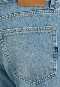 Light blue denim jeans featuring a back pocket with visible stitching, a tan leather patch at the waistband, and a small dark accent detail.