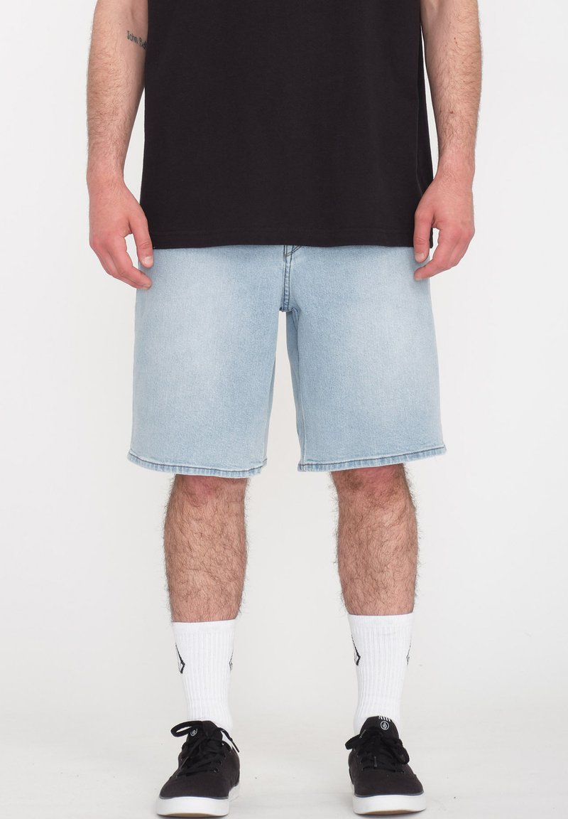 Hellblaue Jeansshorts mit lockerer Passform, knielang, mit rohem Saum, kombiniert mit einem schwarzen T-Shirt und schwarzen Sneakers, ergänzt durch weiße Socken.