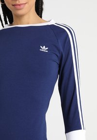 Tummansininen pitkähihaisen paita, jossa on valkoinen kolmiraidan kuvio ja valkoinen kaulus. Rinnassa on pieni valkoinen Adidas-logo. Sujuva kangasrakennus.