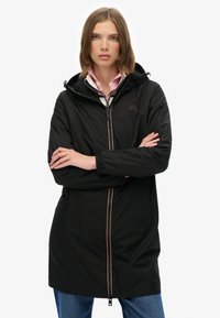 Veste noire imperméable avec capuche, dotée d'une fermeture éclair à l'avant, de deux poches latérales et d'un logo sur la poitrine. Portée par-dessus une chemise à carreaux légers.