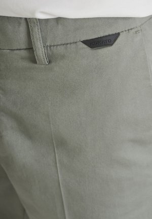 Gros plan sur la poche d'un pantalon vert olive clair avec une étiquette de marque Morato noire et une partie d'une chemise blanche.