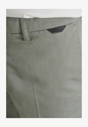 Gros plan sur la poche d'un pantalon vert olive clair avec une étiquette de marque Morato noire et une partie d'une chemise blanche.