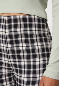 Gros plan d'une personne portant un pantalon à carreaux noir et blanc et un haut à manches longues côtelé vert clair, montrant la taille et une main reposant sur le côté.