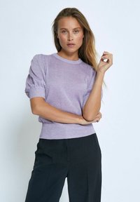 Ljuslila kortärmad tröja med en texturerad, glittrande yta och ribbade manschetter. Stylad med svarta byxor, modellen poserar med armarna i kors.