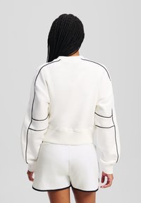 Felpa bianca in cotone con dettagli in piping neri, lunghezza cropped e polsini a coste. Abbinata a pantaloni corti coordinati per un look armonioso.