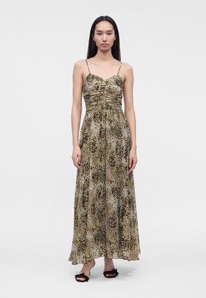 Femme debout portant une robe longue sans manches à imprimé léopard avec fines bretelles et sandales noires ouvertes à l’avant, sur un fond clair uni.