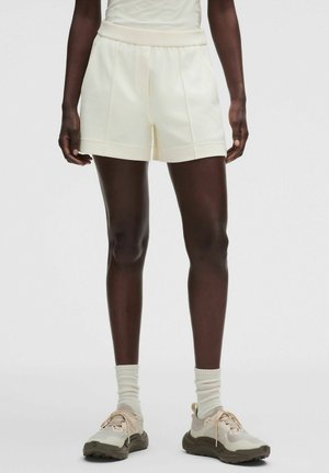 Person iført cremefarvede shorts, hvide sokker og beige og hvide atletiske sko står mod en ensfarvet baggrund.