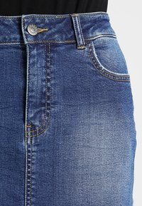 Jupe en denim bleu foncé à taille haute, avec des coutures dorées, des poches avant et une fermeture à bouton métallique à la taille.