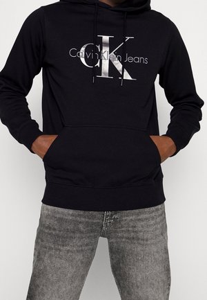 Hoodie in cotone nero con un grande logo argento che riporta la scritta "Calvin Klein Jeans" e una tasca kangaroo frontale, caratterizzato da polsini e orlo a costine.
