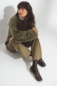 Pair of black flat shoes, square toe design, made of leather. Worn with olive green sweater and suede trousers, against a light background.