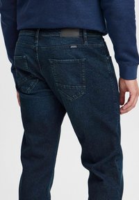 Mörkblå denimjeans med en figurnära passform, med vinklade bakfickor och subtil textur. Läderetikett på midjan för varumärkesidentitet.