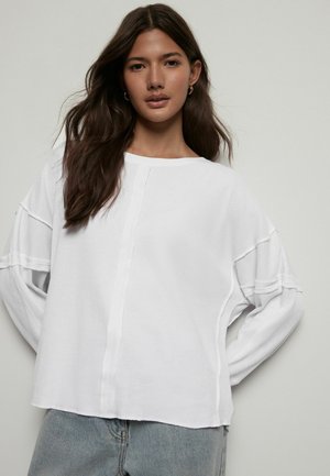 Valge pikka varrukatega shirt, millel on tekstureeritud, kootud muster, lahtine lõige, langevad õlad ja horisontaalsed õmblused.