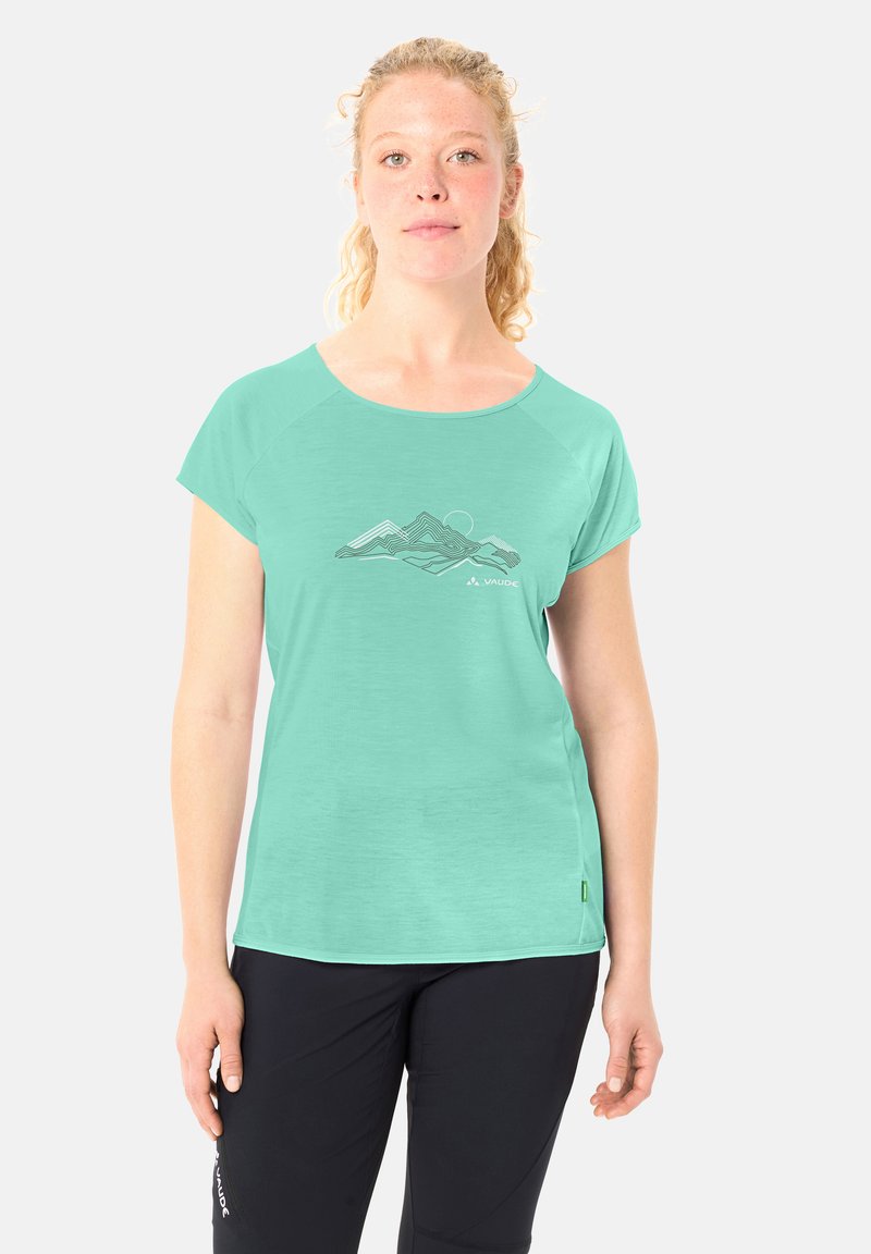 Jeune femme aux cheveux bouclés portant un t-shirt turquoise à manches courtes avec un motif de montagne et un pantalon noir, debout devant un fond blanc.