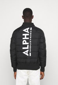Schwarze Steppjacke mit einem gerippten Kragen und Bündchen, ausgestattet mit einem großen weißen "ALPHA"-Logo und dem Text "INDUSTRIES" auf dem Rücken.
