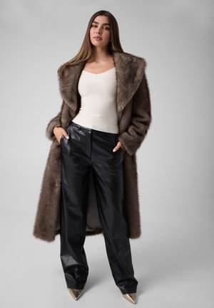 Une femme porte un long manteau de fourrure marron, un haut blanc, un pantalon en cuir noir et des chaussures à talons pointues métallisés, sur un fond de studio gris uni.