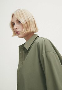 Jeune personne aux cheveux blonds courts portant une chemise oversize vert olive boutonnée, regardant sur le côté sur un fond clair uni.