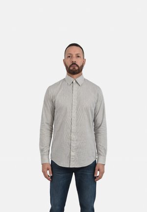 Uomo con capelli corti e barba, indossa una camicia a maniche lunghe grigia e bianca a righe con bottoni e jeans blu scuro, in piedi davanti a uno sfondo bianco.