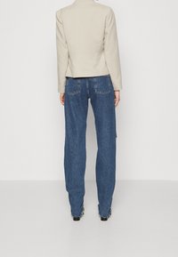 Beżowa kurtka z usztywnionymi ramionami, w zestawieniu z luźnymi niebieskimi dżinsami z tylnymi kieszeniami. Gładka tkanina o minimalnym połysku.