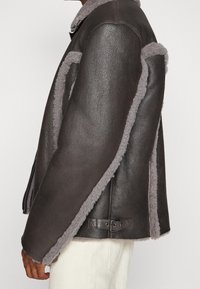 Brun læderjakke med grå fårskindspynt, der har et struktureret design og sidelukningsdetalje med spænde. Glat tekstur, moderne silhuet.