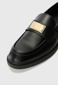Musta nahka loaferit, joissa on kultaisen sävyinen suorakulmainen metallikoriste sangassa ja sileä pinta pyöristetyllä varpaalla.