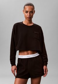 Czarna krótka bluza z długimi rękawami i kieszenią; zestawiona z czarnymi spodenkami z białym paskiem Calvin Klein w talii. Gładka faktura.