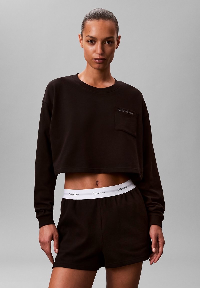 Czarna krótka bluza z długimi rękawami i kieszenią; zestawiona z czarnymi spodenkami z białym paskiem Calvin Klein w talii. Gładka faktura.