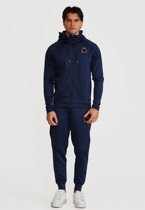 Mann iført marineblå hettejakke med glidelås og matchende joggebukser med gullemblem, stående vendt mot fronten, hvite joggesko synlige.