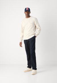Cremefarbener Pullover, marinefarbener Hut mit roten Akzenten, marinefarbene Hose und beigefarbene Sneaker. Lässiges Outfit mit bequemer Passform und strukturierten Materialien.