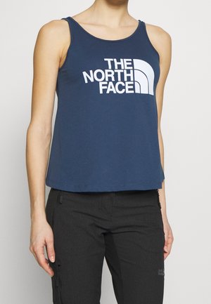 Woman wearing navy blue sleeveless top with "The North Face" logo in white, paired with black pants with zippered pockets.