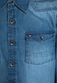 Jasnoniebieska koszula dżinsowa z wygiętym kołnierzykiem, czterema srebrnymi guzikami i kieszenią na piersi z małą czerwoną, białą i niebieską metką.