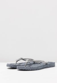Flip-flops med en struktureret grå vævet sål og sølvglitrende remme, der har et minimalistisk design og flad bund for komfort.
