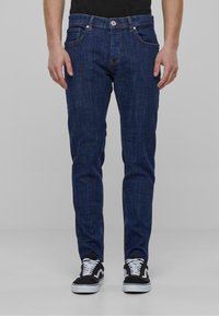 Sötétkék farmernadrág, slim fit szabással, öt zsebbel, gombos záródással és diszkrét varrással. Fekete-fehér sportcipővel párosítva.
