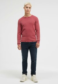 Roter strukturierter Pullover mit langen Ärmeln, Rundhalsausschnitt, kombiniert mit dunkelblauen Jeans und hellen Sneakers. Einfaches, tailliertes Design.