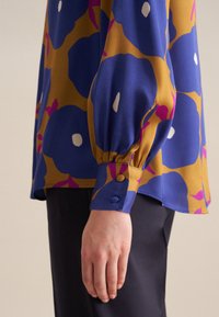 La blusa colorata presenta motivi floreali blu e oro, con un orlo arrotondato e maniche arricciate, fissate da bottoni abbinati sui polsini.