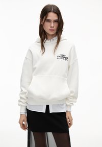 Hoodie blanc avec le texte "TRÈS CHARME EN VRAIE VIE", coupe oversize, poche kangourou, ourlet côtelé, porté par-dessus une chemise bleu clair et une jupe noire.
