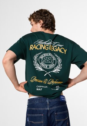 Junger Mann mit lockigem Haar, der ein dunkelgrünes, rennsportinspiriertes T-Shirt und blaue Jeans trägt, zeigt die Rückseite mit Text- und Logodrucken.