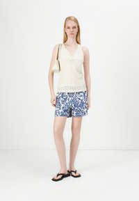 Strikke en kremfarget tank topp med v-hals og knutepunkt, kombinert med mønstrede blå og hvite shorts. Modellen bærer en liten kremfarget håndveske og har på seg svarte flip-flops.