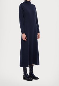Navy Midi-Kleid mit langen Ärmeln, hohem Ausschnitt, lockerer Passform und glatter Textur, kombiniert mit glänzenden schwarzen Knöchelstiefeln.
