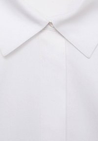 Bílá košile s límečkem a hladkým povrchem, která má zapínání na knofl�íky u krku a čistý, minimalistický design.