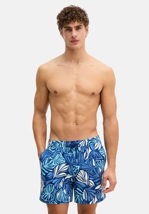 Uomo senza maglietta con capelli ricci che indossa pantaloncini da bagno blu con motivi di foglie tropicali bianche e azzurre, mani in tasca, sfondo neutro.
