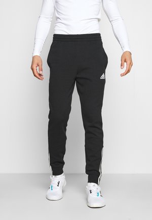 Mann trägt schwarze Adidas-Trainingshose mit weißen Streifen und Logo, steht mit entspannten Armen an den Seiten, trägt weiße Sportschuhe und langärmeliges Oberteil.