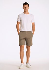 Pantaloni corti in cotone khaki con una vestibilità rilassata, dotati di tasche laterali e di una vita con chiusura a bottone, abbinati a una maglietta bianca semplice e scarpe da ginnastica.