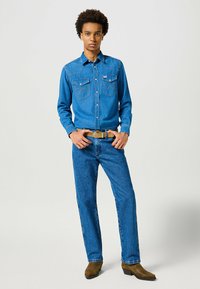 Camicia in denim blu con due tasche sul petto, abbinata a jeans blu e stivali marroni. Presenta una vestibilità classica e dettagli di cucitura.