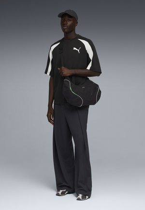 Schwarzer Mann trägt ein schwarz-weißes Puma-Shirt, weite dunkle Hose, Mütze, Turnschuhe und hält eine schwarze Puma-Schultertasche, steht vor grauem Hintergrund.